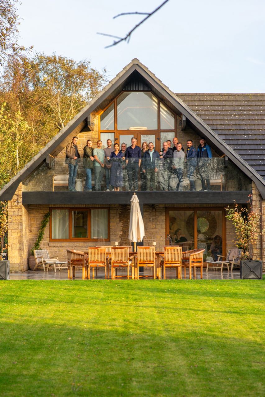 A large group of guests gathered on the balcony of Warren House at golden hour, with the gardens in front